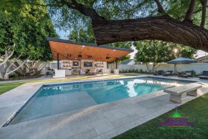 Custom backyard landscape with large covered patio with fireplace and outdoor kitchen with large trees and steps to custom swimming pool
