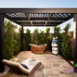 Custom backyard design with patterned metal shade structure, outdoor tub and privacy hedges.