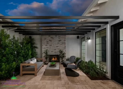Customized backyard design with slatted metal shade structure, floor-to-ceiling brick fireplace and privacy hedges.